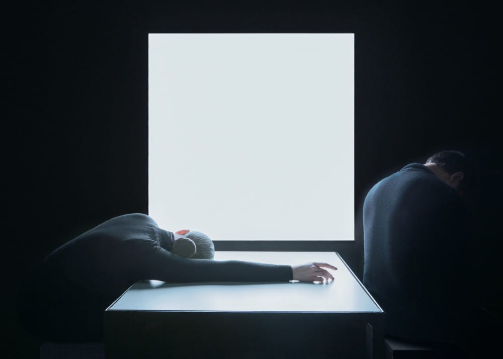 un homme assis et une femme affalée  sur une table avec un écran lumineux de couleur blanche en fond 