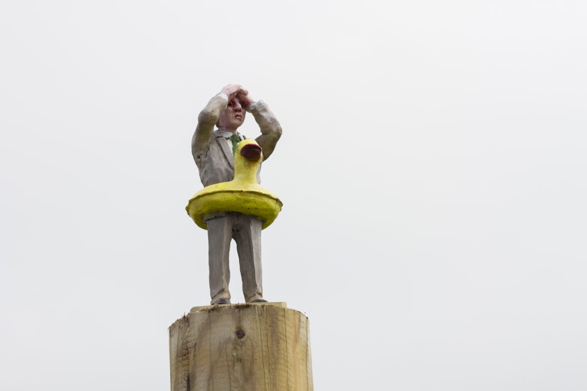 Isaac Cordal, un personage miniature portant une bouée en forme de canard au sommet d'un poteau. 