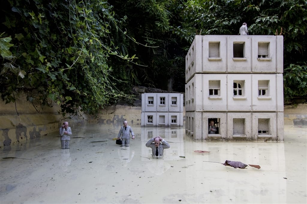 Isaac Cordal, Plusieurs humains miniatures dans un bassin blanc avec des bâtiments engloutis. 