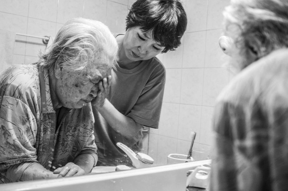 Photo en noir et blanc représentant une femme âgée se faisant rincer le visage par une jeune femme. 