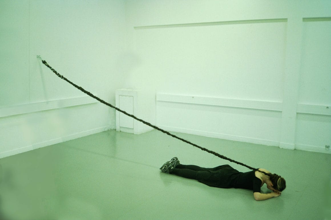 Reine Louve allongée sur le sol, une longue tresse accrochée au mur, performance, 