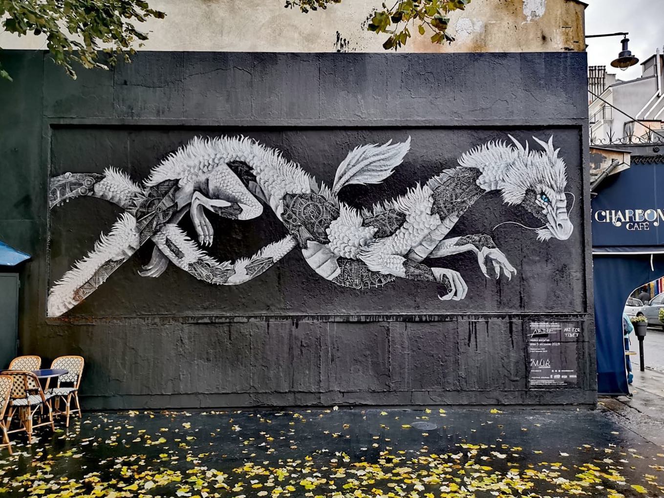 Sur un mur peint en noir, un dragon en gris et blanc dont on voit les rouages en dessous de ses écailles.