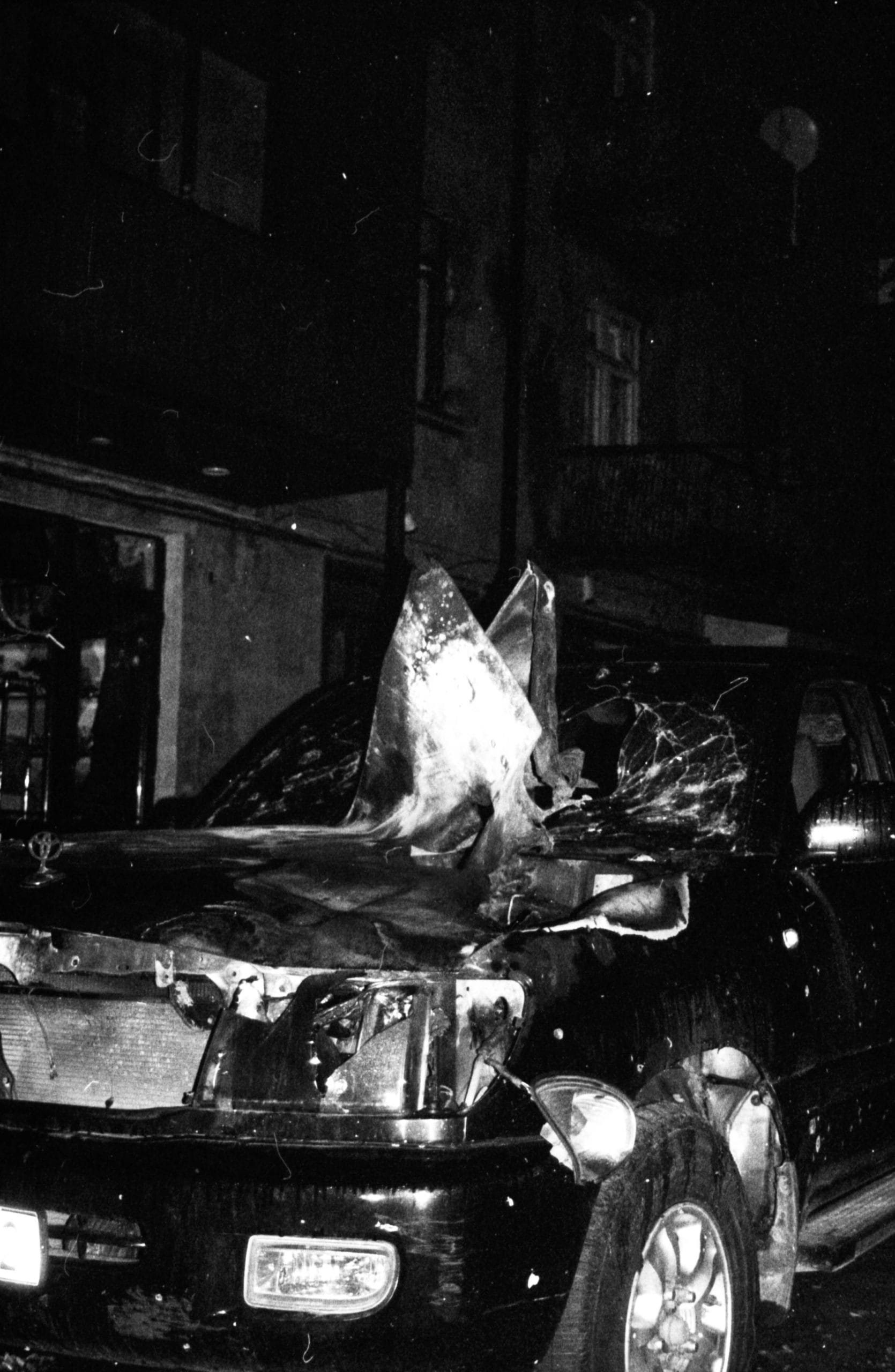 Photo en noir et blanc, très obscure, mis à part l'avant qui est illuminée : il s'agit d'une voiture détruite qui a probablement été touchée par un projectile