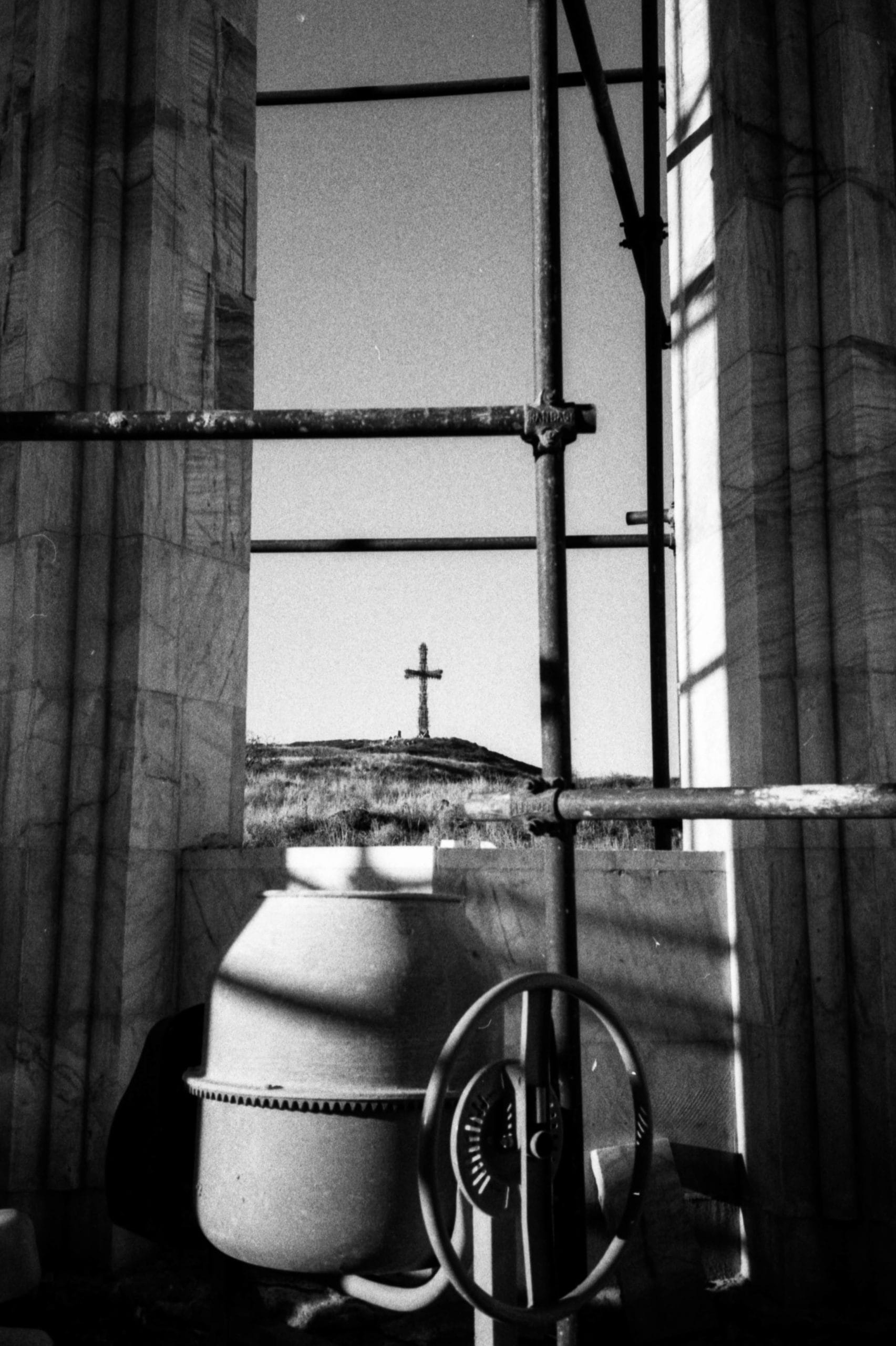 Photographie en noir et blanc d'une fenêtre par laquelle on aperçoit une croix sur une colline 