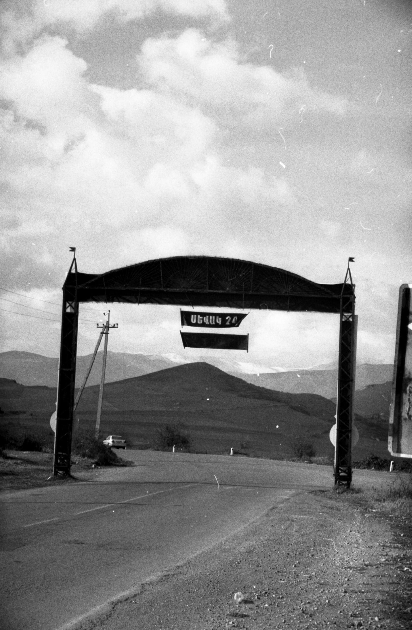 Photographie en noir et blanc d'une route au-dessus de laquelle est un écriteau.