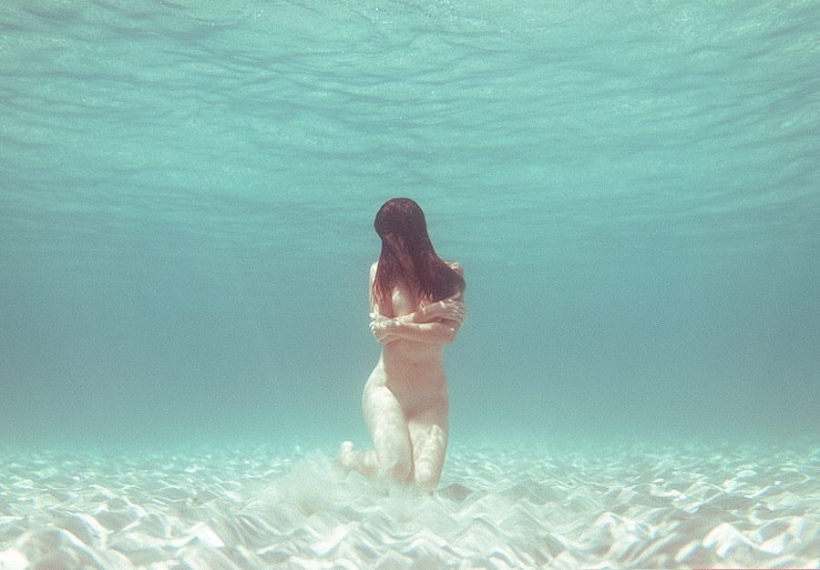 Une femme est à genou sur le sable sous l'eau. Elle serre ses bras contre sa poitrine et ses cheveux cachent son visage.