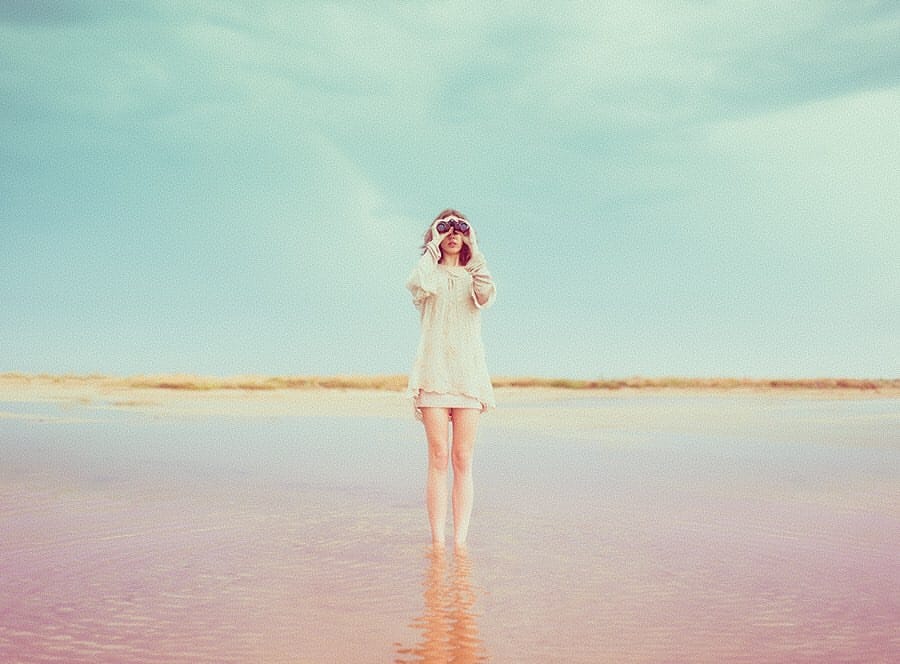 Une femme se tient droit devant l'objectif, les pieds dans la mer. Elle observe quelque chose au loin avec ses jumelles