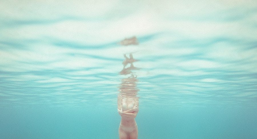 Photographie prise sous l'eau, on voit une femme nue serrer ses bras contre sa poitrine