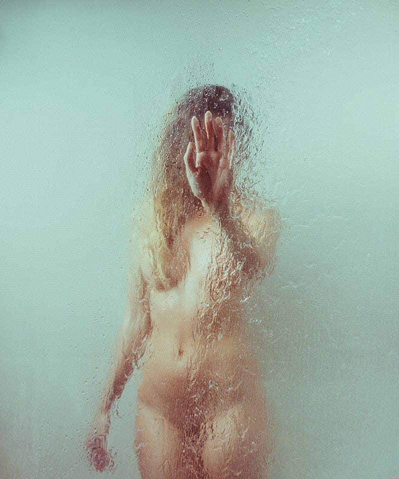 Photographie d'une femme nue derrière une vitre couverte d'eau. Elle a sa main posée sur 
