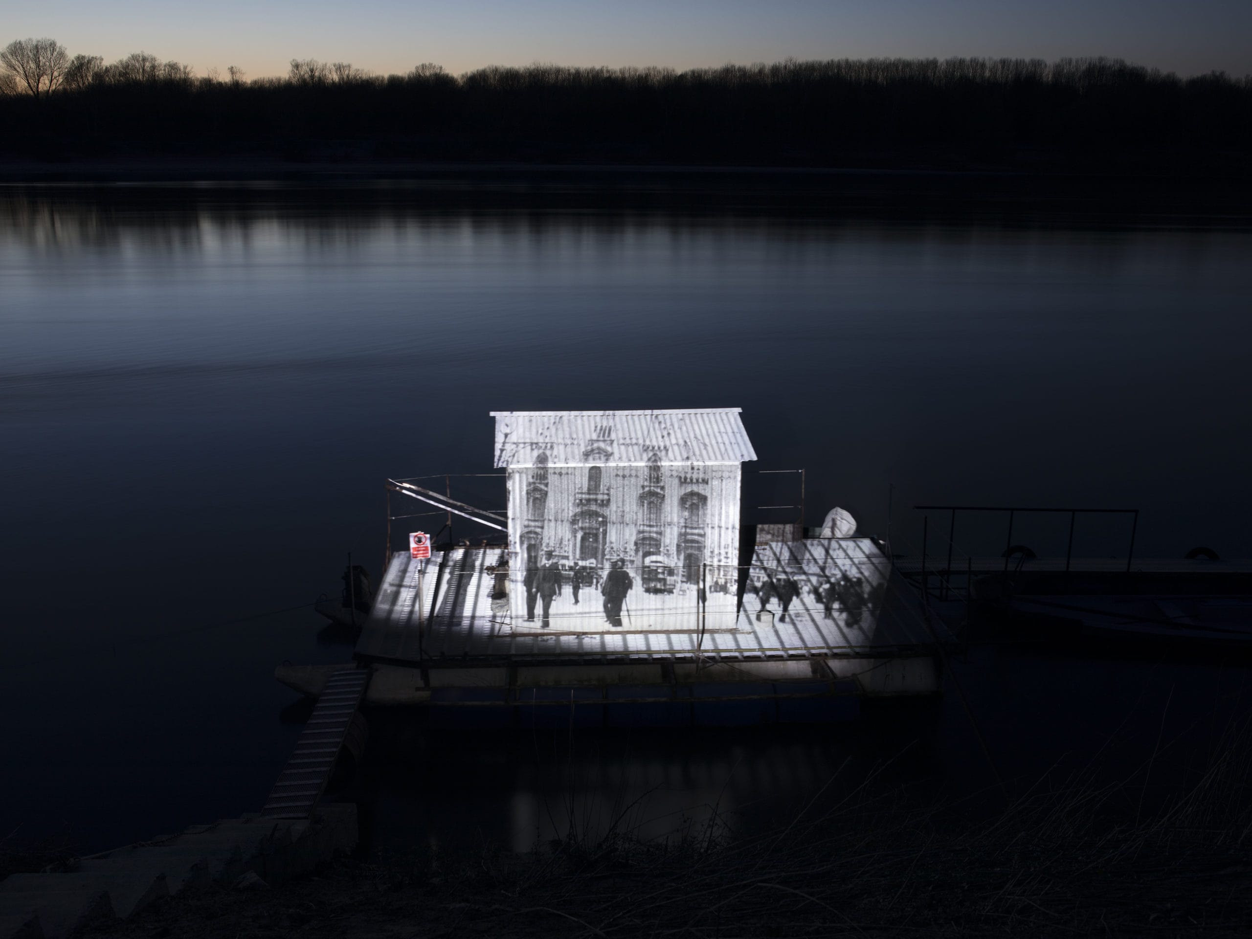 Photographie d'un fleuve de nuit, une maison en bois sur l'eau sur laquelle est projetée une photo