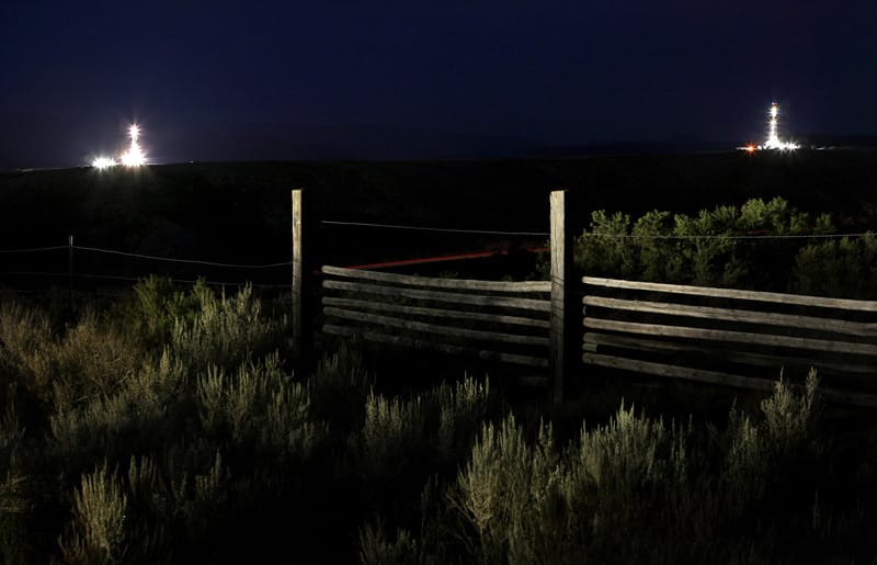 Une clôture dans la nuit. Végétation très présente. Au loin, deux forages dont de la lumière dans cette atmosphère sombre.