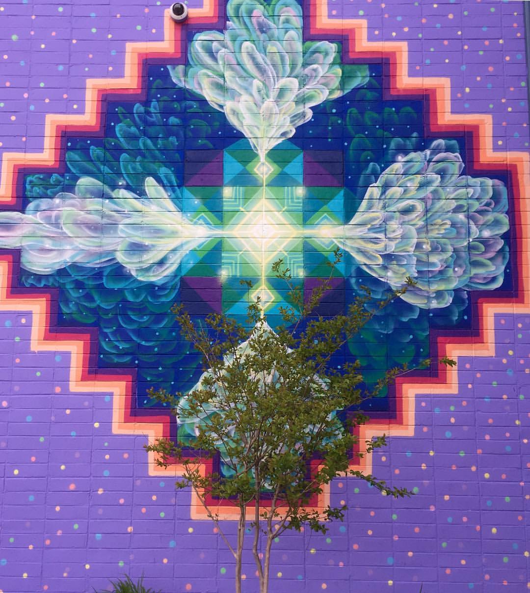 Motif géométrique et symétrique (bleu, violet, vert et rose) sur un mur, un arbre est devant