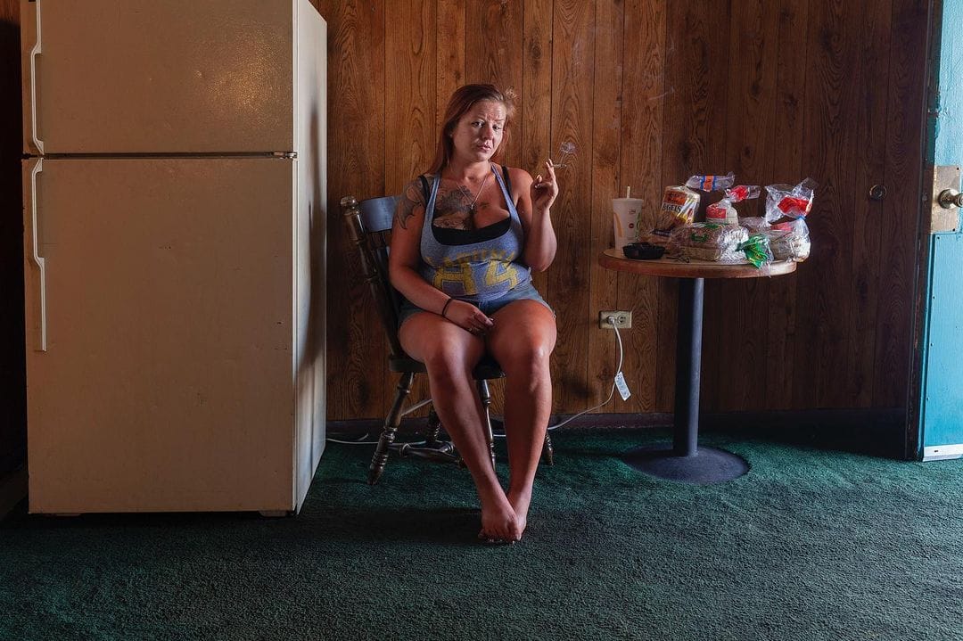 Une femme assise sur une chaise entre un frigo et une table sur laquelle il y a des paquets de pain de mie. Elle fume une cigarette.