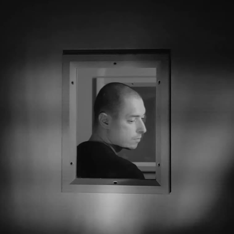 Photographie en noir et blanc d'un homme, à travers la fenêtre d'une porte insonorisée.