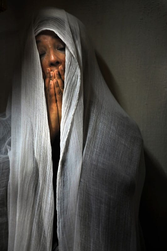 Une femme prie avec les mains devant le visage, sous un linge blanc.