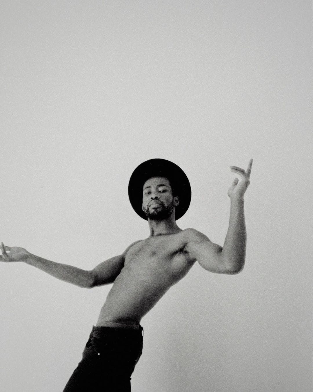 Autoportrait du chanteur Annael. Torse nu, il danse, le corps en mouvement. Il porte un chapeau noir qui contraste avec le fond blanc. 