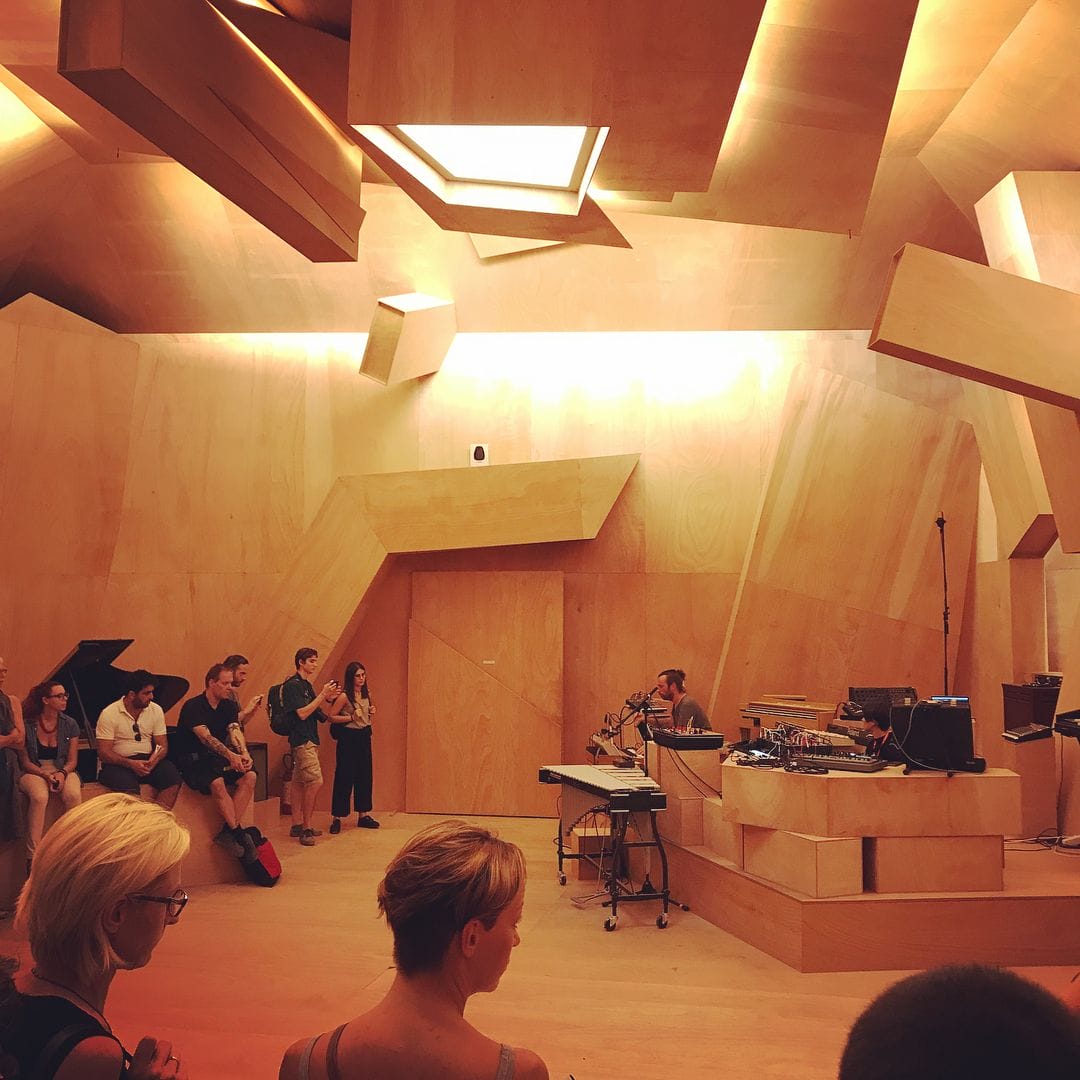 Photographie de l'installation. Grande salle recouverte de bois taillés de manière géométrique, avec un public qui observe un musicien, dans le coin, entouré d'instruments.