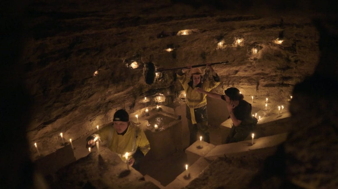 Dans une grotte, Myd tient la perche au dessus d'un point d'eau, pendant que le second personnage lui indique le geste qu'il doit faire et que le troisième allume des bougies.
