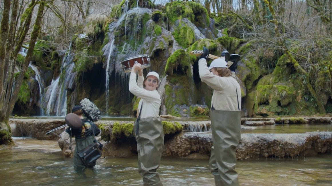 Les trois personnages traversent une rivière, en tenant leurs instruments au dessus de leurs têtes , paysage sauvage.