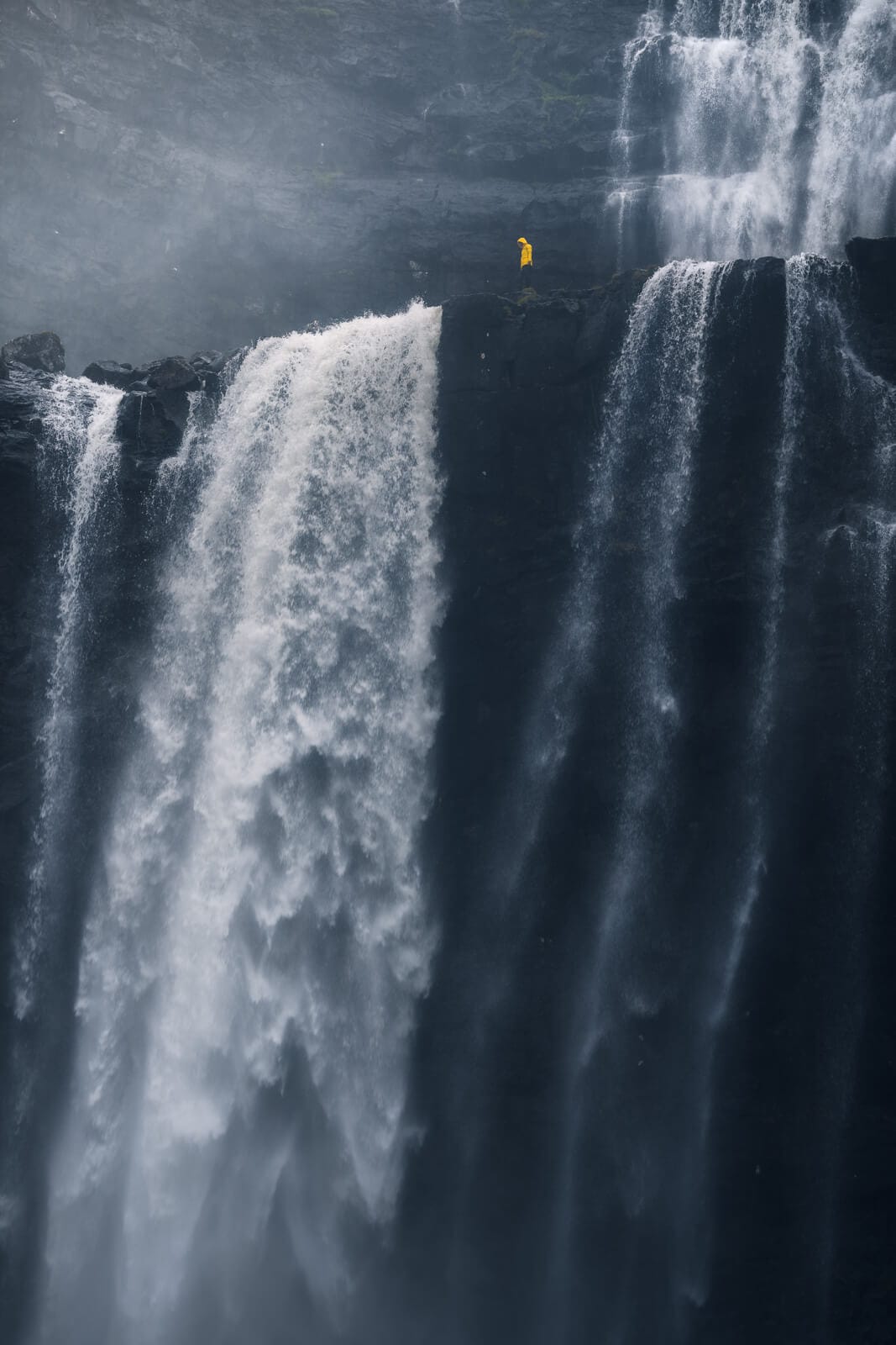 Plan très gris, bord de falaise et chutes d'eau à écume blanche. Une personne se tient au bord, avec un ciré jaune dont la couleur ressort face au roches.