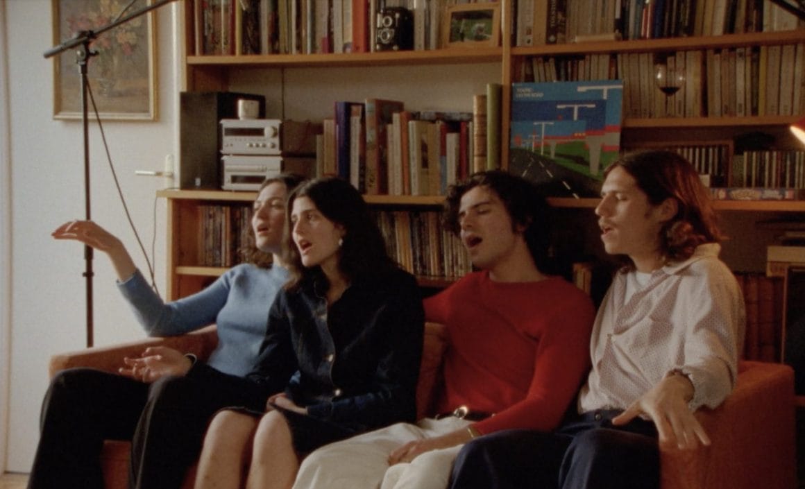 Plan sur les quatre choristes, assis sur un canapé devant une grande bibliothèque. Deux filles à droite, deux garçons à gauche, chantent le refrain en se concentrant.