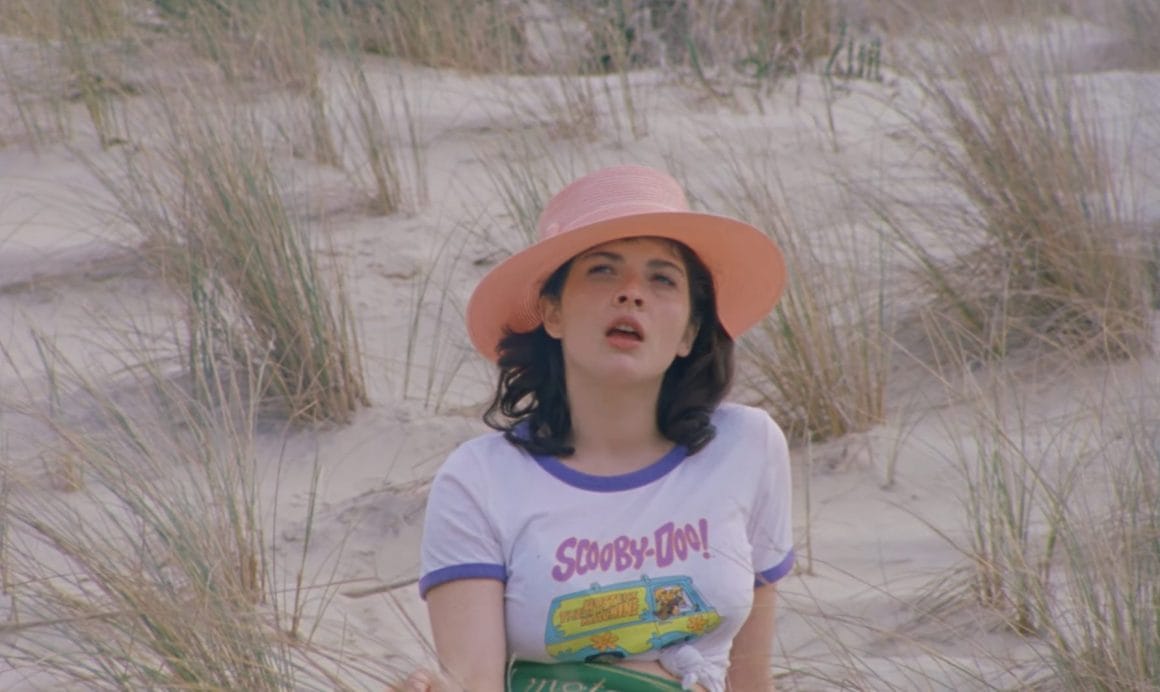 La chanteuse est assise sur une dune de sable, un grand chapeau rose sur la tête/ Elle regarde l'horizon en chantant.