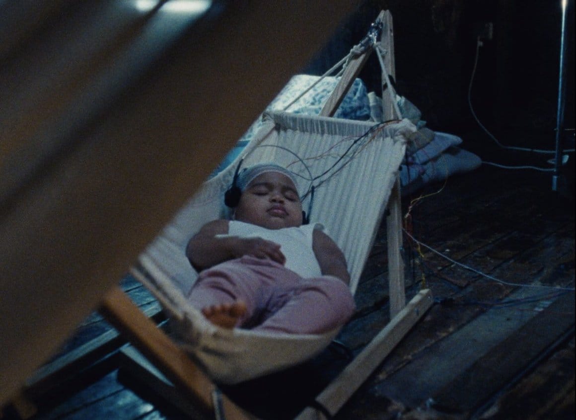 Bébé endormi sur un petit hamac posé sur le sol, casque audio sur les oreilles. Des câbles sont posés sur son crânes et le relient à d'autres éléments, hors champs.