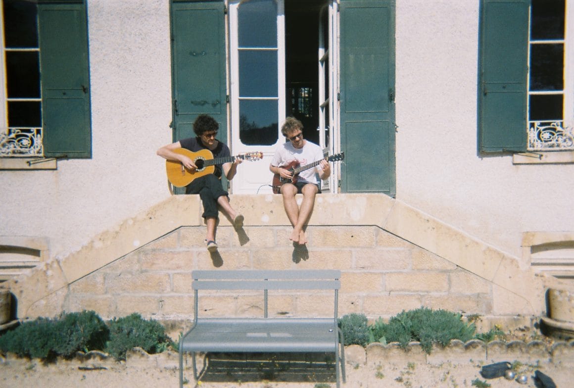 Les deux musiciens sont assis sur le bord d'un escalier d'une grande maison, baignée de soleil. Tous les deux jouent de leurs guitares, les lunettes de soleil sur le nez.