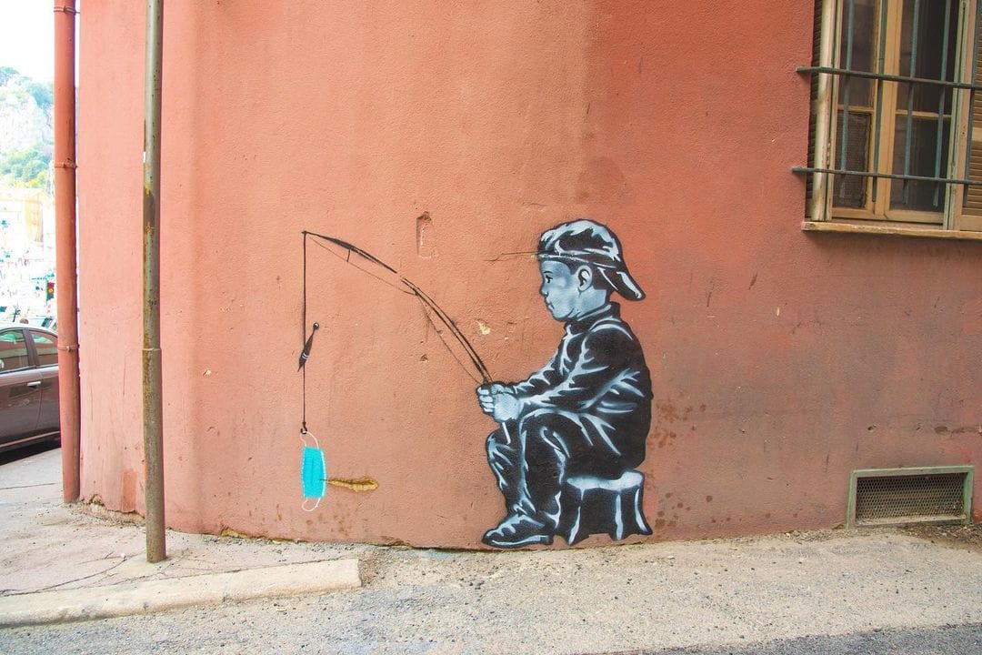 Peinture en noir et blanc au mur. Petit garçon qui tient une cane à pêche dans ses mains. En couleur, au bout du fil, un masque chirurgical.
