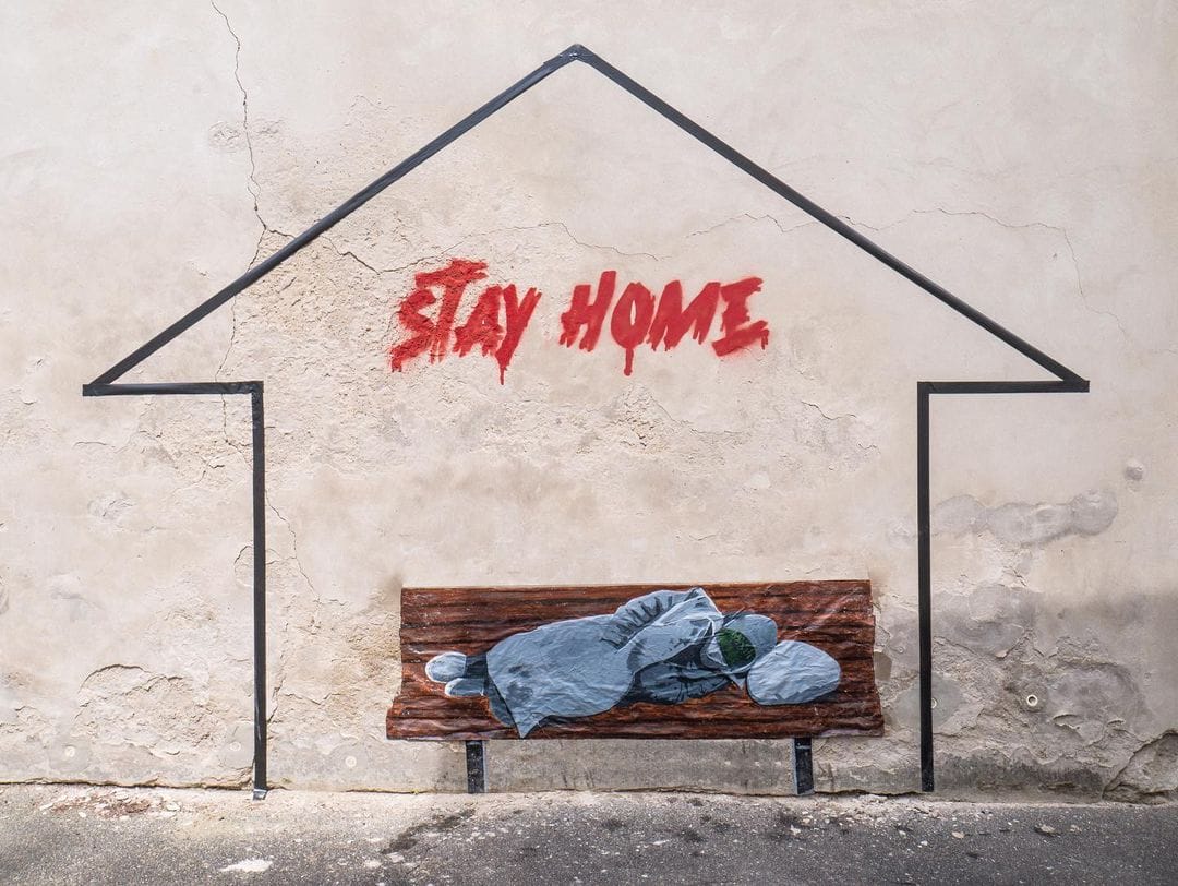 Pan de mur, silhouette grossière de maison, au dessus d'un banc peint, sur lequel une personne dort. Au dessus, en rouge, l'inscription "STAY HOME". 