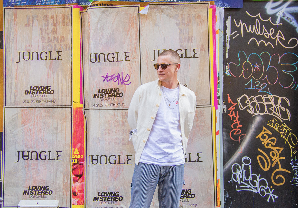 Tom McFarland pose devant un mur où sont placardées des affiches du nouvel album de Jungle.