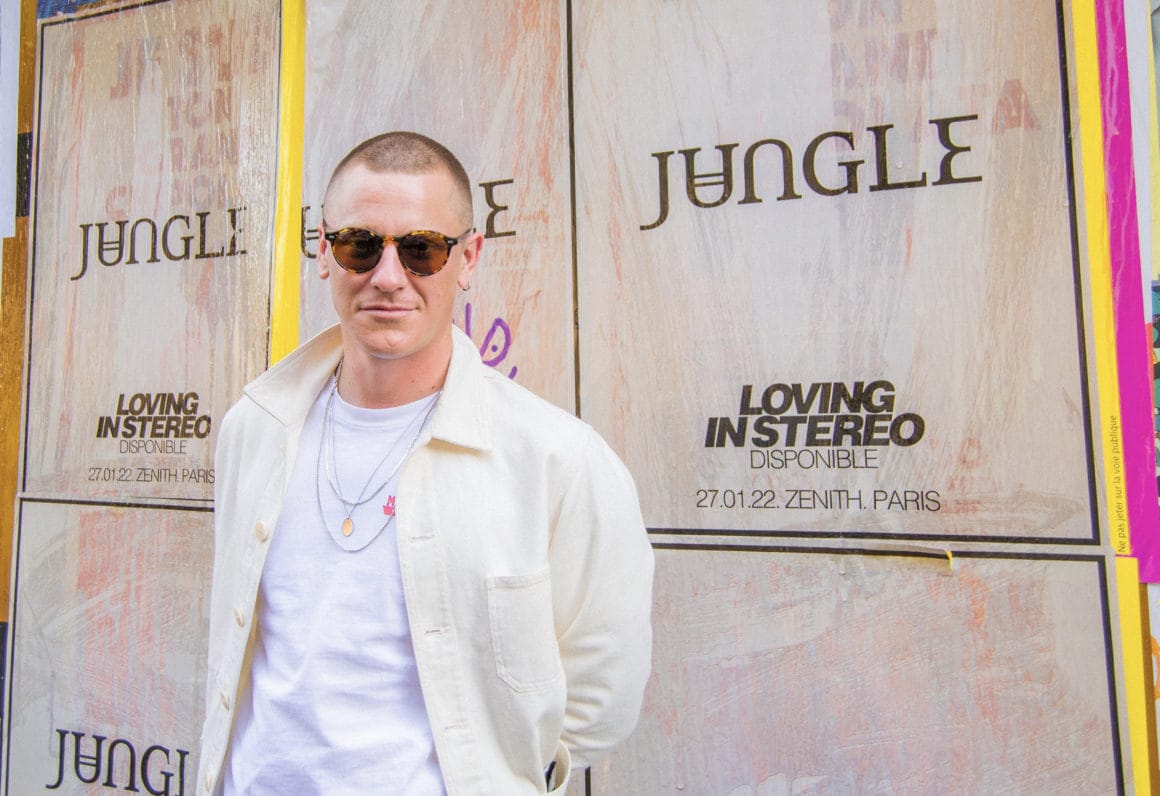 Tom McFarland pose devant un mur où sont placardées des affiches du nouvel album de Jungle (format portrait).
