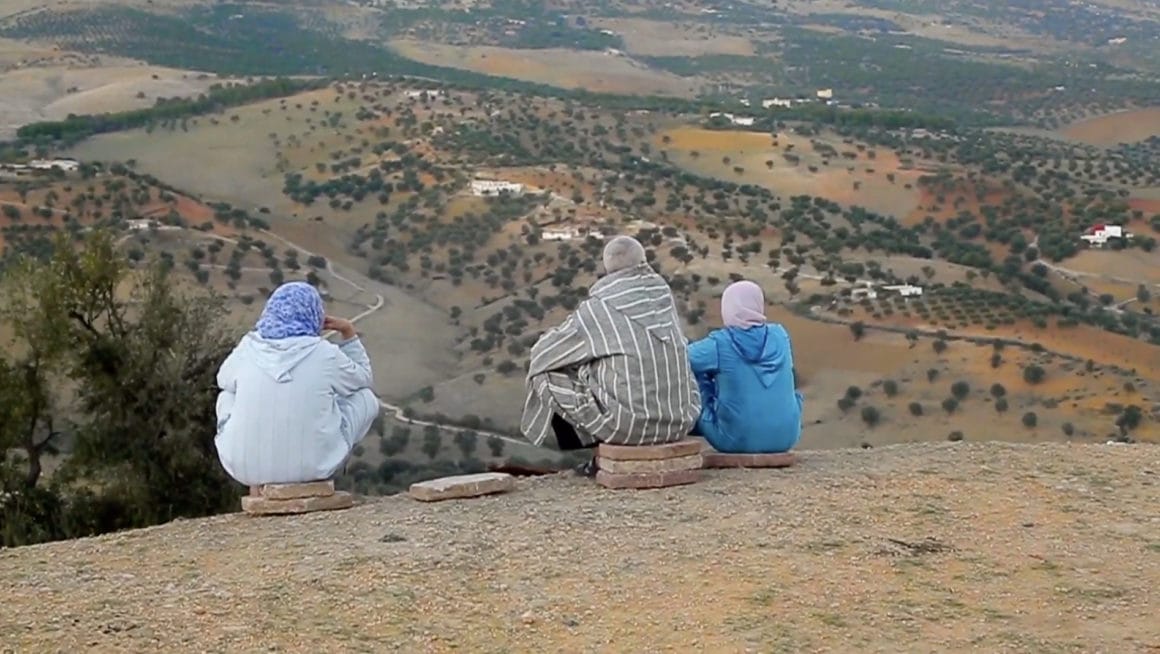Extrait de la vidéo "La vie de tous les jours". Deux femme set un homme sont assis, de dos, à même le sol d'une colline surplombant un désert parsemé de points verts.