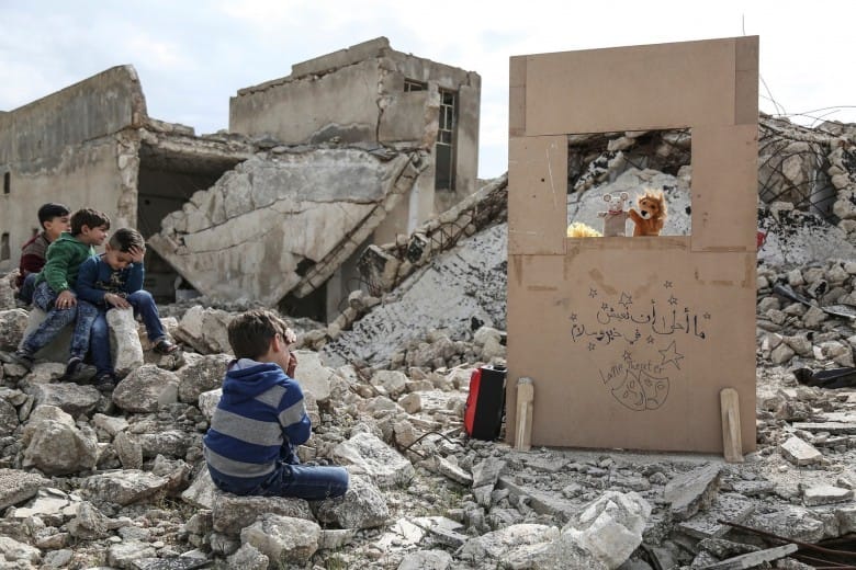 Des enfants sont assis dans les décombres d'un village en Syrie. Devant eux, un théâtre de marionnettes en carton.