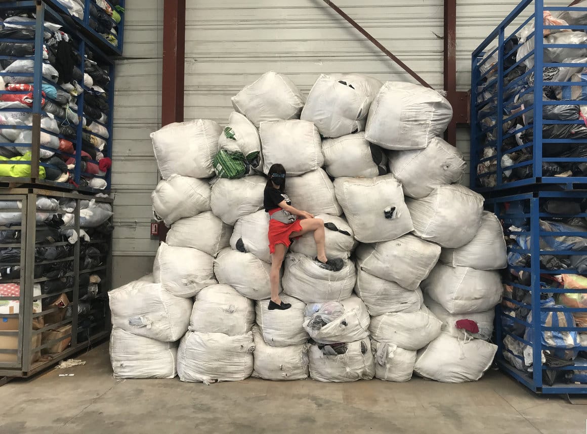 Photographie à l'intérieur d'un entrepôt Le Ralis. Une femme pose devant une pile colossale de sacs de vêtements.