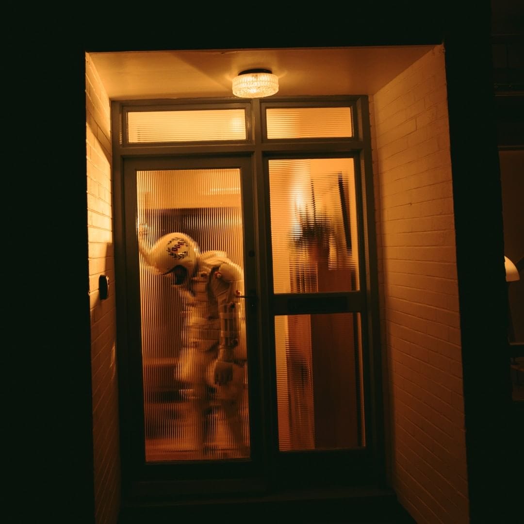 Un cosmonaute se tient, visiblement épuisé, dans un hall d'immeuble. On l'aperçoit de manière déformé, à travers la vitre de la porte. La lumière crée un effet de clair obscur.