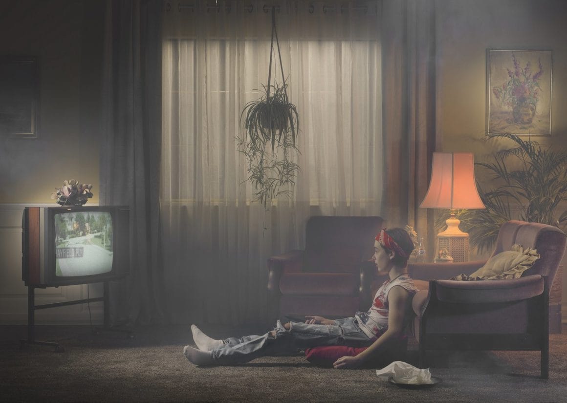 Jeune homme assis devant télévision dans salon rideaux et lampe allumée