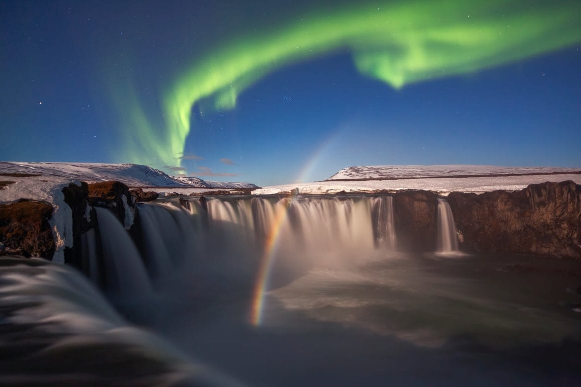 Cette photo représente une aurore boréale prise en Islande, au dessus d'une grosse cascade.