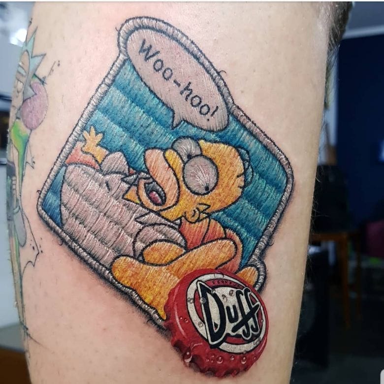 Ce tatouage de Duda Lozano représente Homer Simpson assis sur son canapé essayant d'attraper une capsule de bière Duff.