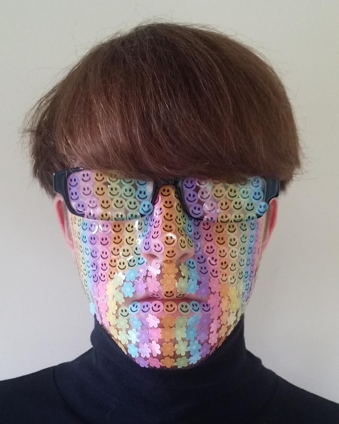 L'artiste Adam Hillman pose avec sa coupe au bol et ses lunettes, la tête recouverte de petites gommettes colorées.