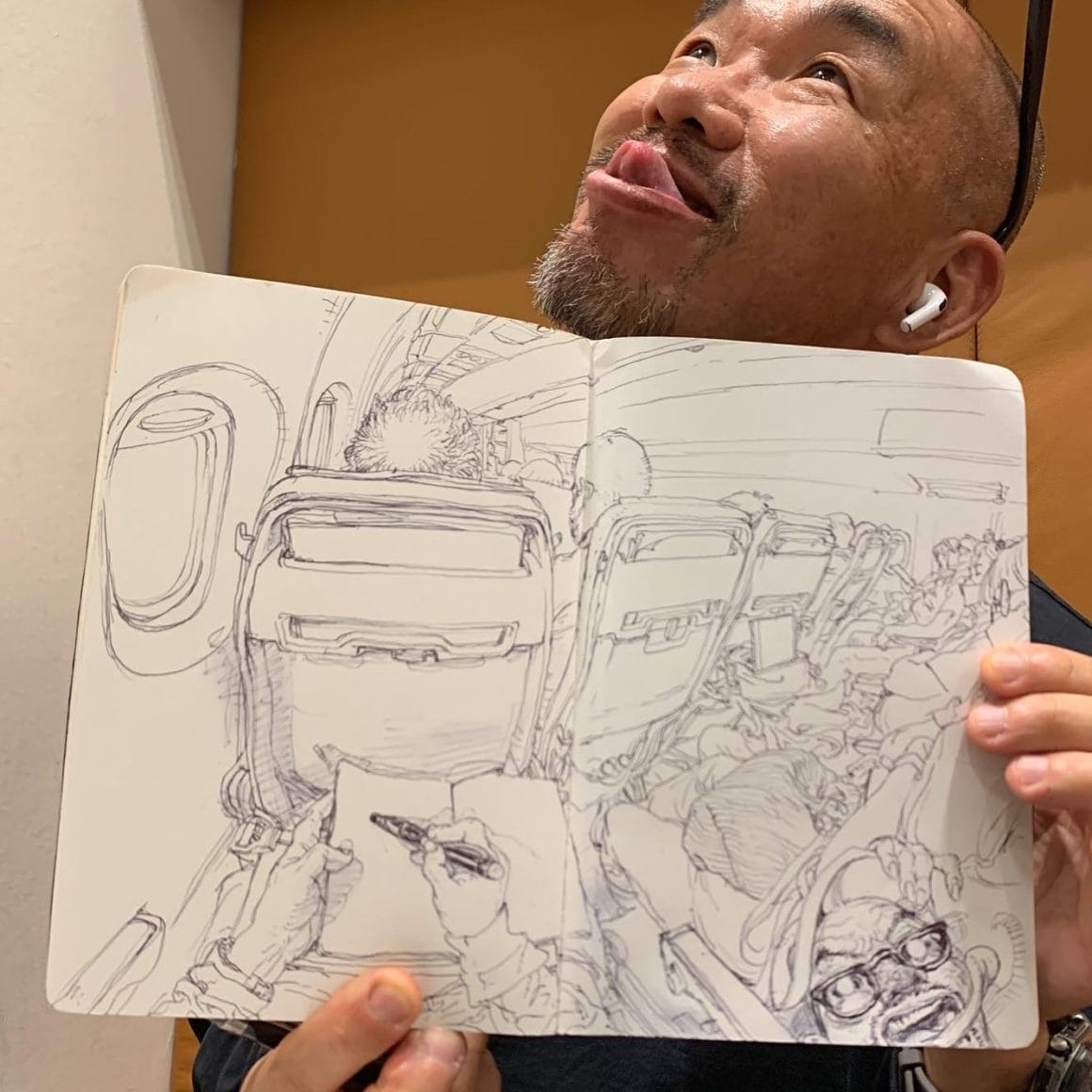 Kim Jung Gi se prend en photo avec son dessin dans les main. Il représente ses mains en train d'elles même dessiner dans l'avion. Nous sommes à la place de ses yeux. Dans le coin de la feuille en baas à droite, il y a une caricature de lui