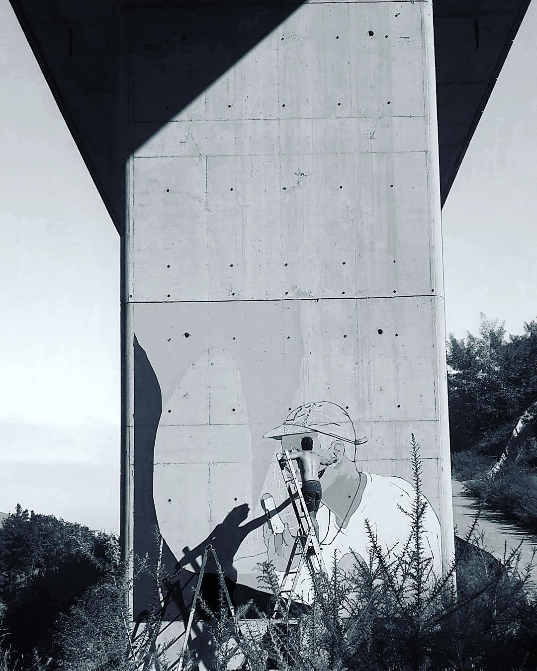 L'artiste Mr.Sor2 peint sa nouvelle fresque en dessous d'un pont. La photo est en noir et blanc alors l'ombre de l'artiste paraît faire partie de l'oeuvre 
