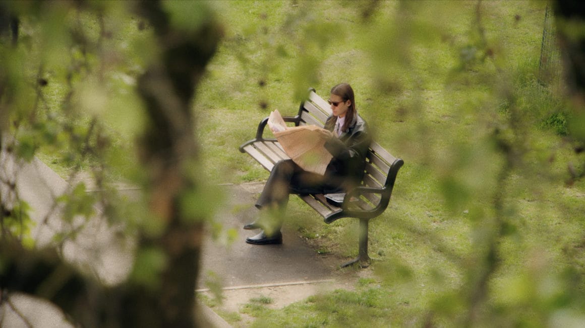 Photographie de Saint DX au coeur de la nature. Il lit un journal sur un banc entouré de verdure.