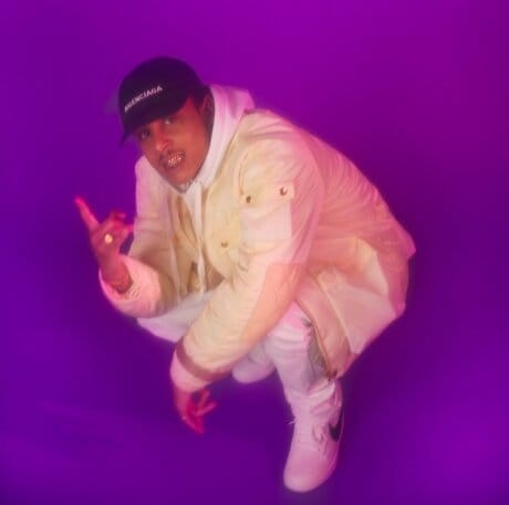Photo où l'on voit Ateyaba avec une veste blanche, une casquette, des bagues et des grillz. Il est accroupis et fait un signe devant un fond violet.