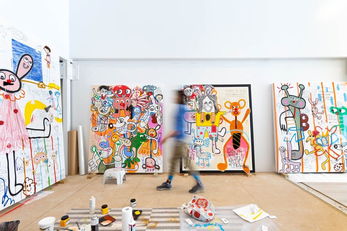 Galerie d'art avec plusieurs tableaux colorés et un atelier avec du matériel. Un homme marche au milieu, il est flou 