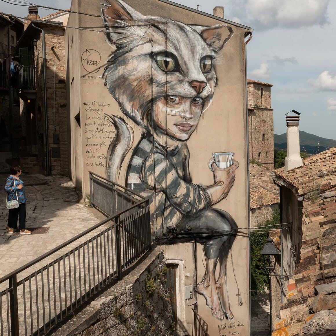 Fresque réaliser par le duo Herakut. Une enfant avec une queue de chat et une tête de chat qu'elle porte comme une capuche est assise et tient un verre de lait dans sa main droite.