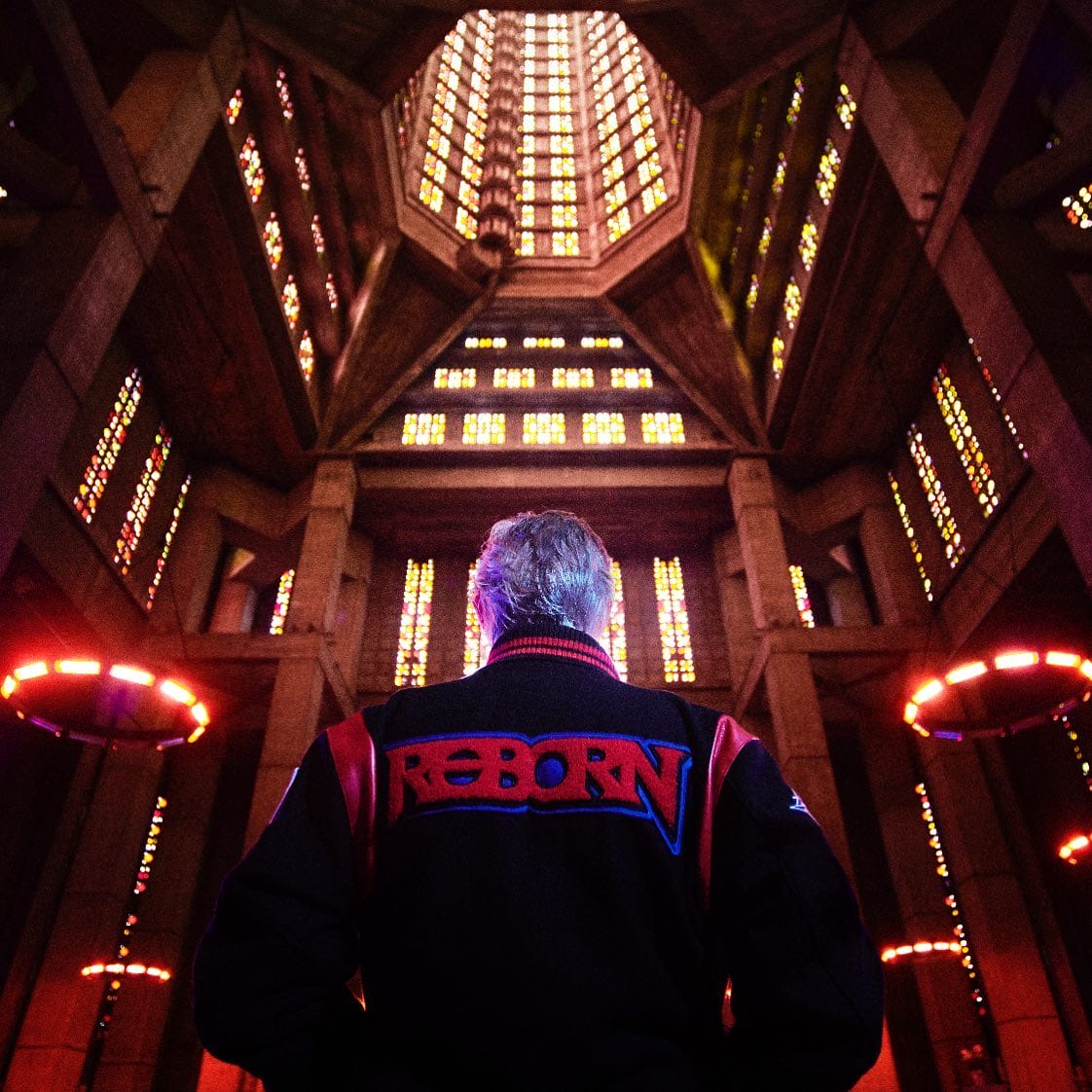 Kavinsky, vu de dos dans une veste « Reborn », se tient au centre d'un bâtiment éclairé par des lumières orangées.