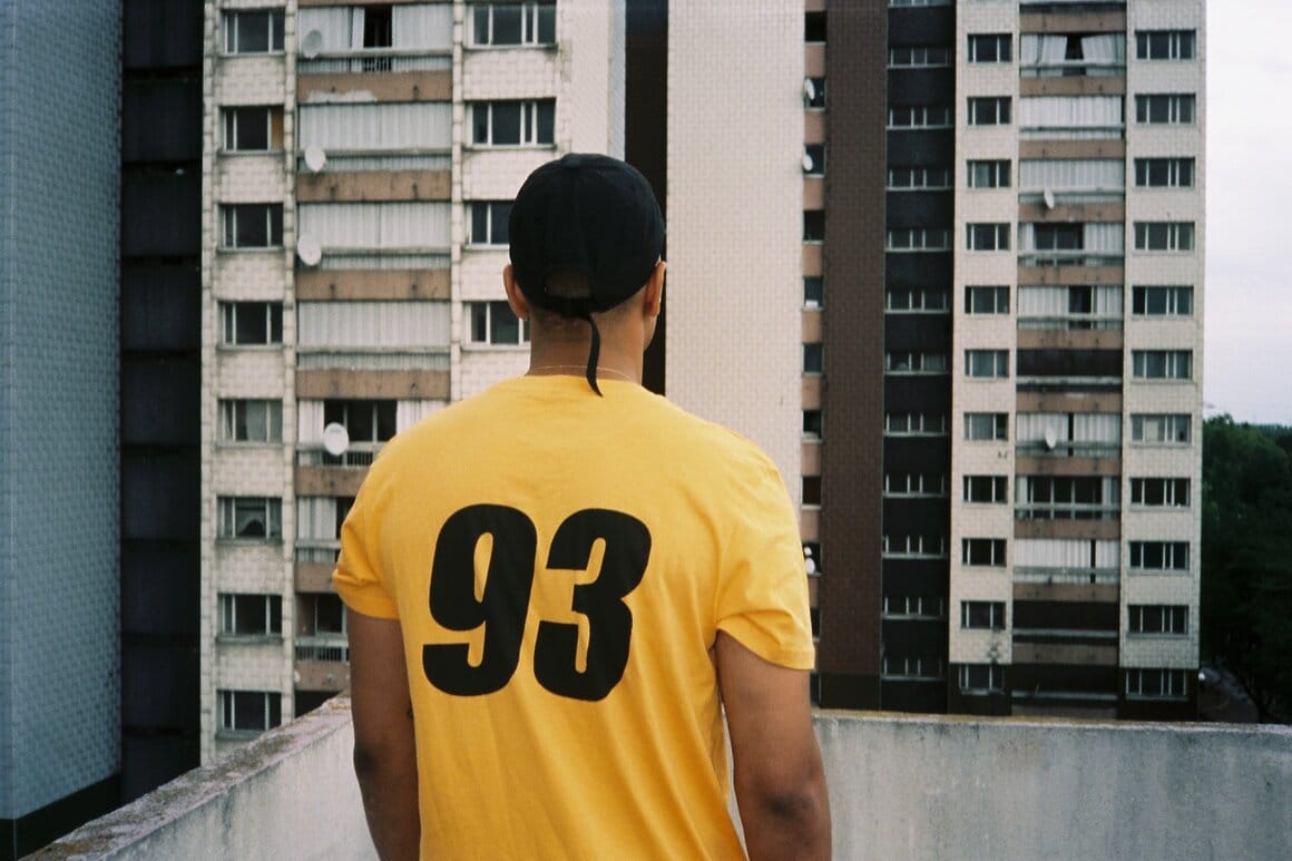 Un homme portant un tee-shirt jaune marqué « 93 » dans le dos regarde les immeubles d’un grand ensemble.