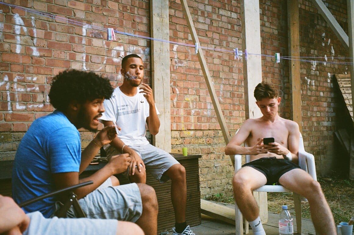 Trois jeunes hommes sont assis devant un mur de briques, l’un fume tandis qu’un autre regarde son téléphone.