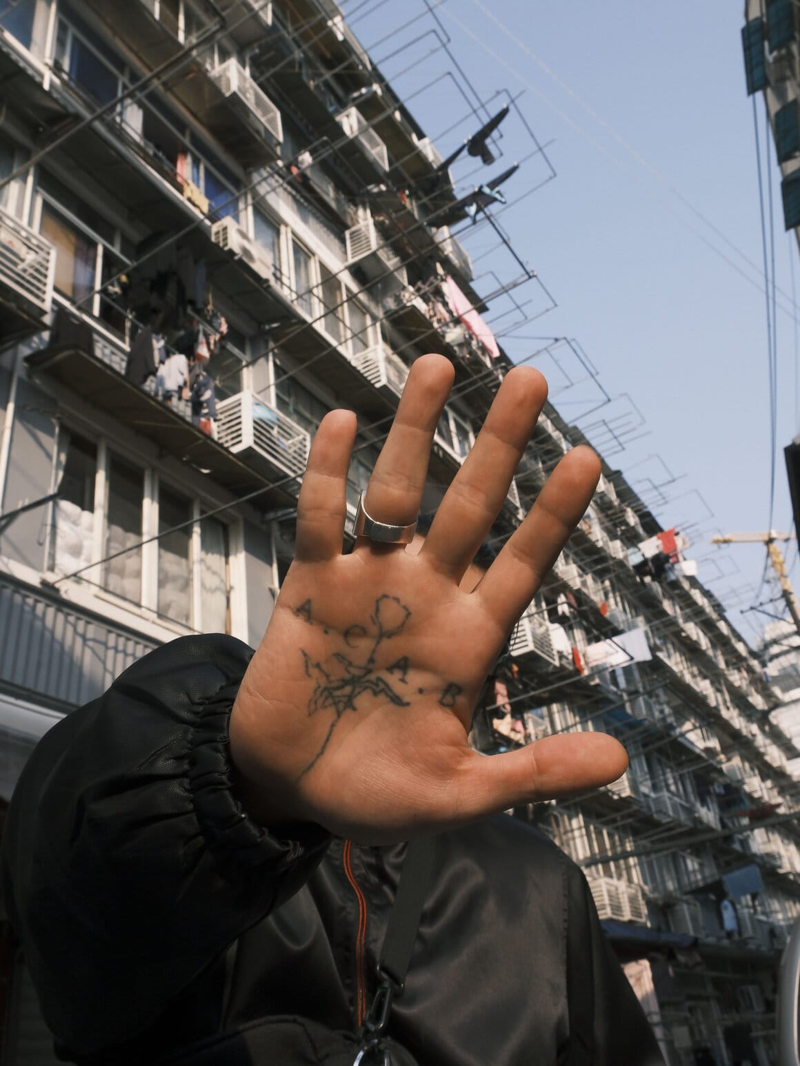 Une main tatouée est tendue vers l’objectif devant la façade d’un immeuble où du linge sèche aux fenêtres.